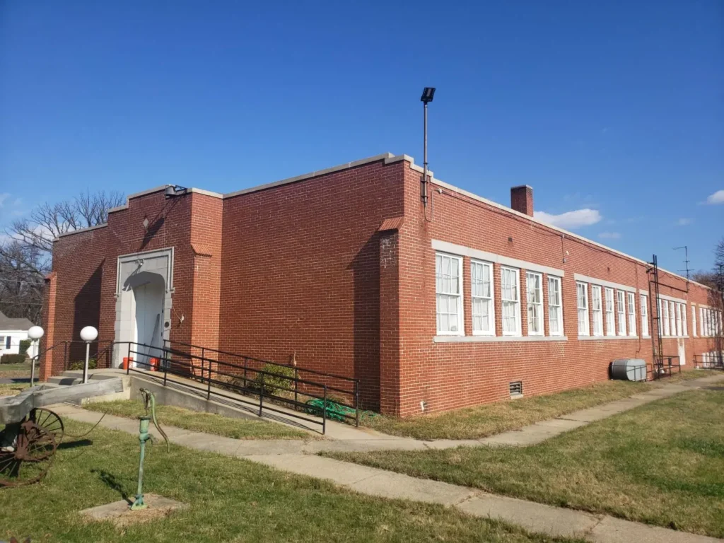The outside of an old brick 1-story building with a white door and windows down the length of the structure.